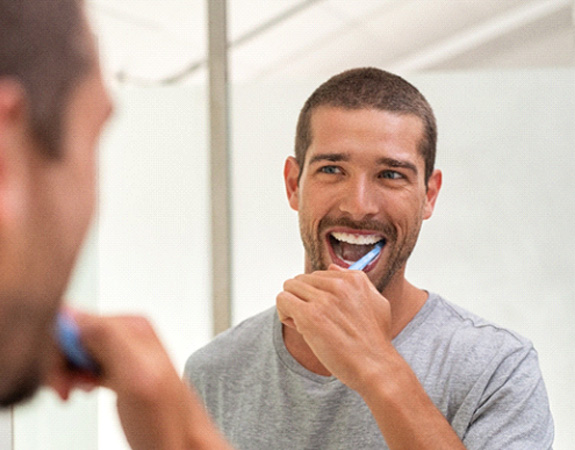 A smiling man brushing his teeth in front of a bathroom mirror